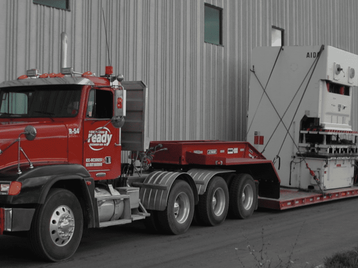 A large red truck is parked next to a building, featuring a prominent grille and multiple lights on the roof. The truck is towing a flatbed trailer that carries a sizable industrial machine, secured with straps. The background consists of a gray metal wall and large windows, indicating a commercial or industrial setting. The truck's design includes a bold "ready" logo and a decorative striped pattern on the front bumper.