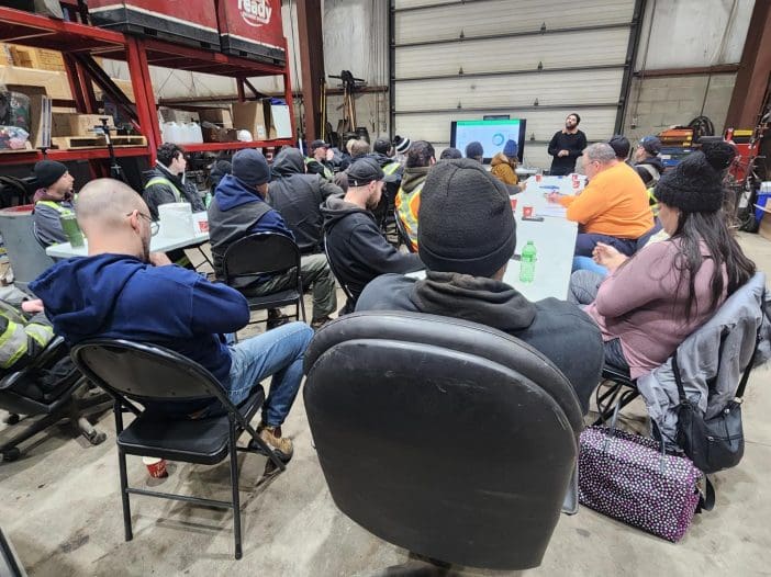 A group of people is seated in a large industrial space, attending a meeting or presentation. The attendees are mostly dressed in work attire, with some wearing safety vests and winter hats. In the foreground, several chairs are visible, with a focus on the back of one chair. A screen at the front displays information, while a speaker stands beside it, engaging the audience. The environment features metal beams and storage containers in the background, typical of a warehouse setting.