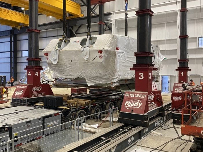 A large industrial setup features a heavy-duty lifting system with four vertical columns, each equipped with hoisting mechanisms. A large object, covered in protective wrapping, is suspended from the lifting apparatus. The structure is located in a spacious warehouse with high ceilings, and there are various pieces of equipment and materials visible on the floor, including a flatbed trailer and a powered lift. The lifting system is branded with "READY" and has a capacity of 600 tons.