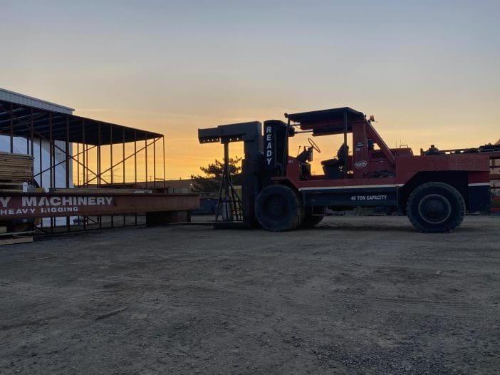 A large red forklift with a capacity of 40 tons is positioned on a gravel surface. It features a raised fork at the front and is parked next to a steel beam labeled "HEAVY MACHINERY." In the background, a partially constructed building is visible, with wooden pallets stacked nearby. The sky is illuminated with warm hues of orange and yellow, indicating either sunrise or sunset.