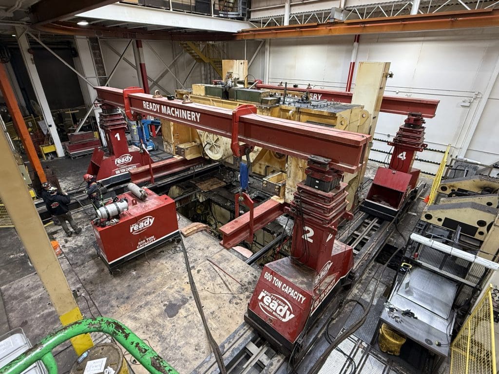 A large industrial machinery setup is visible, featuring multiple red hoisting systems labeled with numbers. The machinery is positioned on a track, with two prominent hoists on either side, each capable of lifting 600 tons. Surrounding the machinery are various tools and equipment, including a winch and several boxes. Two workers in safety gear are present, engaged in the operation. The environment appears to be a workshop or industrial facility, with overhead structures and a mix of machinery components in the background.
