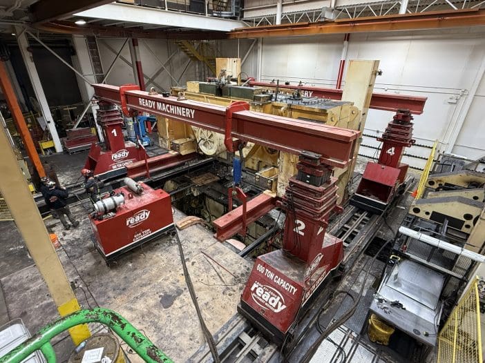 A large industrial machinery setup is visible, featuring multiple red hoisting systems labeled with numbers. The machinery is positioned on a track, with two prominent hoists on either side, each capable of lifting 600 tons. Surrounding the machinery are various tools and equipment, including a winch and several boxes. Two workers in safety gear are present, engaged in the operation. The environment appears to be a workshop or industrial facility, with overhead structures and a mix of machinery components in the background.