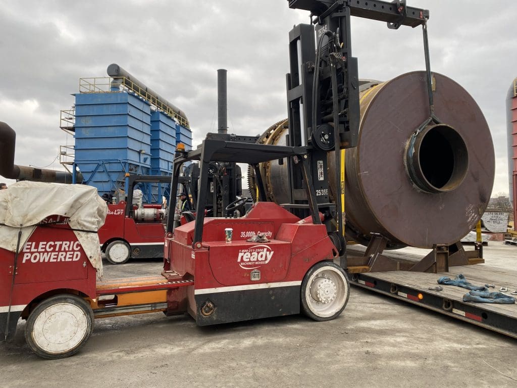 A red electric forklift is positioned next to a large cylindrical metal object, which is being lifted by the forklift's attachment. The background features a blue industrial structure with multiple sections, along with other machinery and equipment. The scene is set outdoors under a cloudy sky, indicating an industrial environment. The forklift has a label indicating its electric power source and a capacity of 35,000 pounds.
