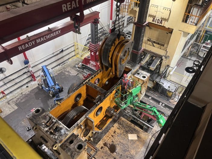 A large industrial workspace features heavy machinery and equipment. In the center, a partially disassembled machine with large gears is visible. A worker in safety gear operates a green lift, positioned near the machine. Above, a red overhead crane labeled "READY MACHINERY" is seen, with additional machinery and tools in the background. The floor is marked with wear, indicating heavy use.