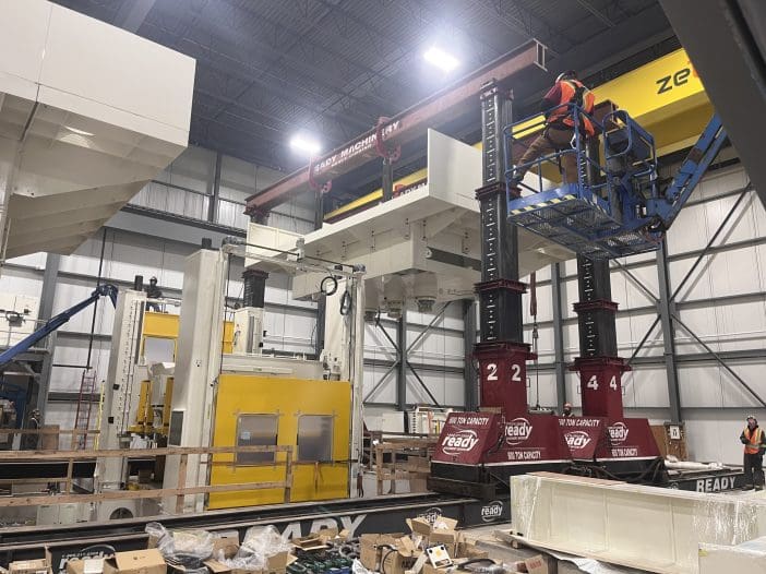 A large industrial space features heavy machinery and equipment. In the center, a significant piece of machinery is being lifted by two large red hydraulic jacks, each labeled with a capacity of 600 tons. Above, a crane is positioned to assist in the lifting process. Workers in safety vests are present, one on a lift platform and others observing the operation. Surrounding the machinery are various tools, boxes, and materials, indicating an active work environment. The walls are made of metal panels, and the ceiling is high, typical of a warehouse or manufacturing facility.