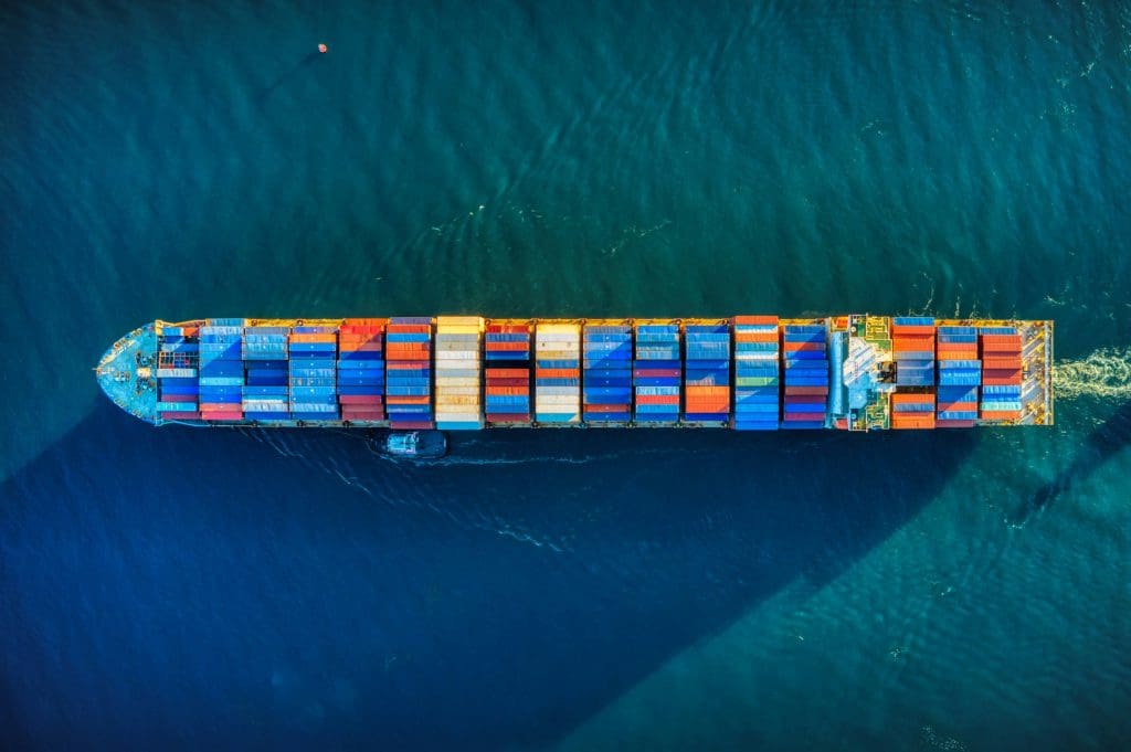 A large cargo ship loaded with multicolored shipping containers is sailing on deep blue water, viewed from directly above. The containers are arranged in neat rows, predominantly in blue, red, orange, and white colors. A small boat is positioned alongside the cargo ship near the front. The ship's wake is visible behind it.