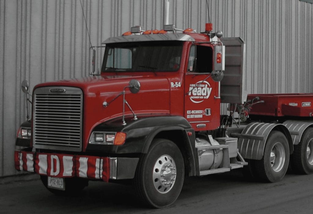 Red semi-truck cab with black fenders and silver wheels, featuring the "Ready" company logo on the door. The truck has a large front grille, multiple orange lights on the roof, and a striped red and white bumper guard. The background is a gray industrial building.