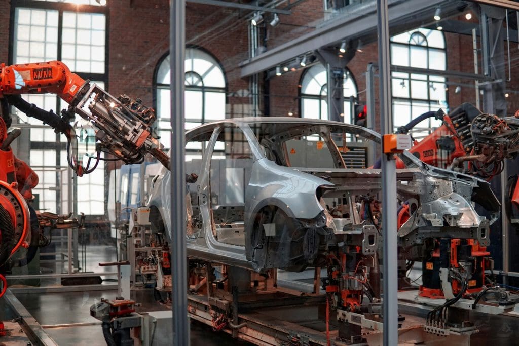 A partially assembled silver car body is positioned on an assembly line inside a factory. Several orange robotic arms labeled "KUKA" are working on the car, likely performing welding or assembly tasks. The factory has large arched windows and industrial lighting, with a spacious and clean environment.