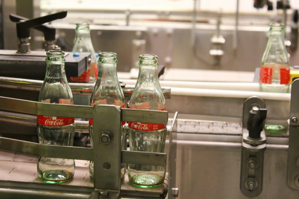 Empty glass Coca-Cola bottles with red labels are positioned on a conveyor belt in a bottling or packaging facility. The bottles are held in place by metal guides as they move along the production line. The background shows industrial equipment and machinery.