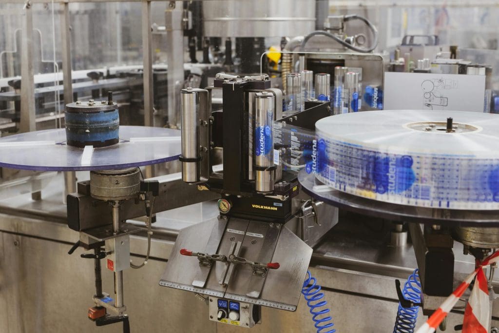 A labeling machine in a production line, with a roll of transparent labels featuring blue text and graphics on the right side, and several cylindrical metal containers positioned for labeling in the center. The machine has various mechanical components, including rollers, clamps, and control knobs, with a gauge and the brand name "VOLKMANN" visible on the front. The background shows a blurred industrial setting with more machinery and equipment.