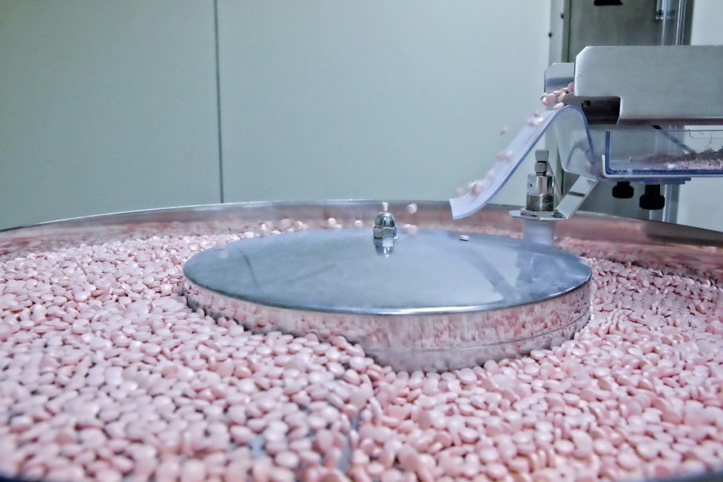 A pharmaceutical machine sorting or processing numerous small, round, pink tablets on a large circular metal tray with a central rotating component. Tablets are being dispensed onto the tray from a chute on the right side.