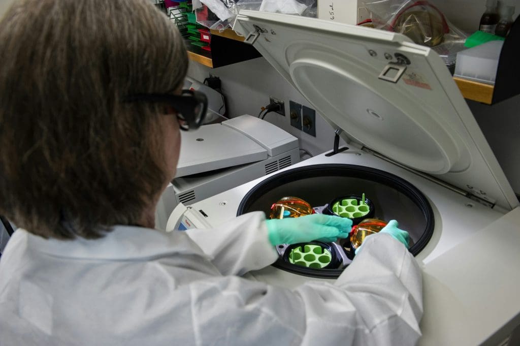 A person wearing a white lab coat, green gloves, and protective glasses is operating a centrifuge machine in a laboratory. The centrifuge lid is open, revealing rotor buckets with orange covers and green tube holders inside. The background includes lab equipment and shelves with various supplies.