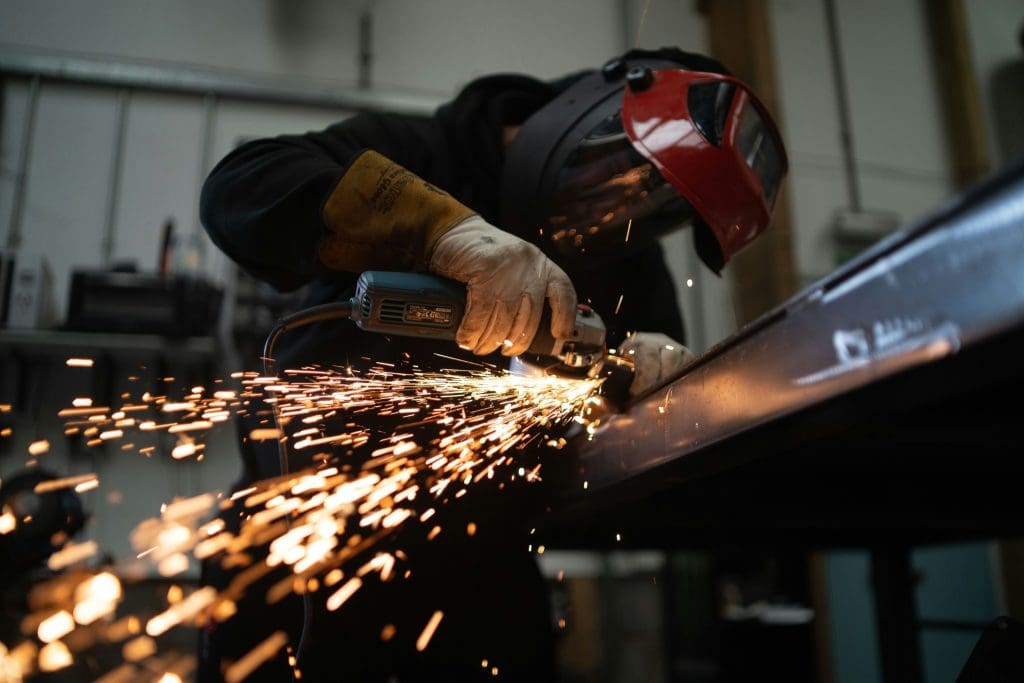 A person wearing protective gloves and a red welding helmet is using an angle grinder to cut or grind a metal beam, producing bright sparks in a workshop setting.