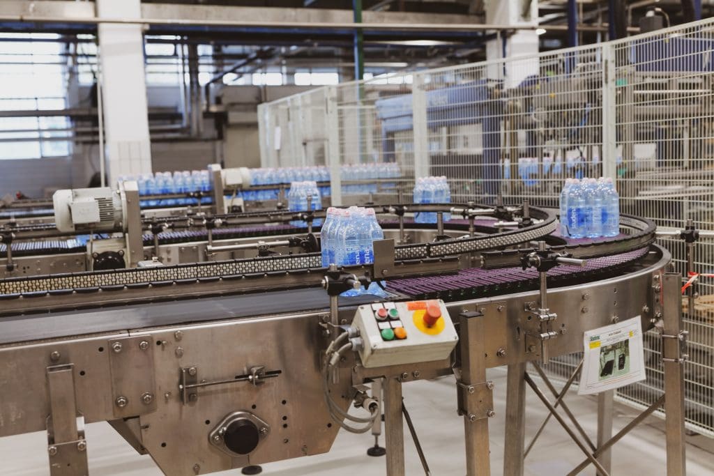 A stainless steel conveyor system in a factory setting transporting multiple packs of bottled water. The conveyor curves and has a control panel with buttons and an emergency stop switch attached to it. The background shows industrial equipment and a fenced-off area.