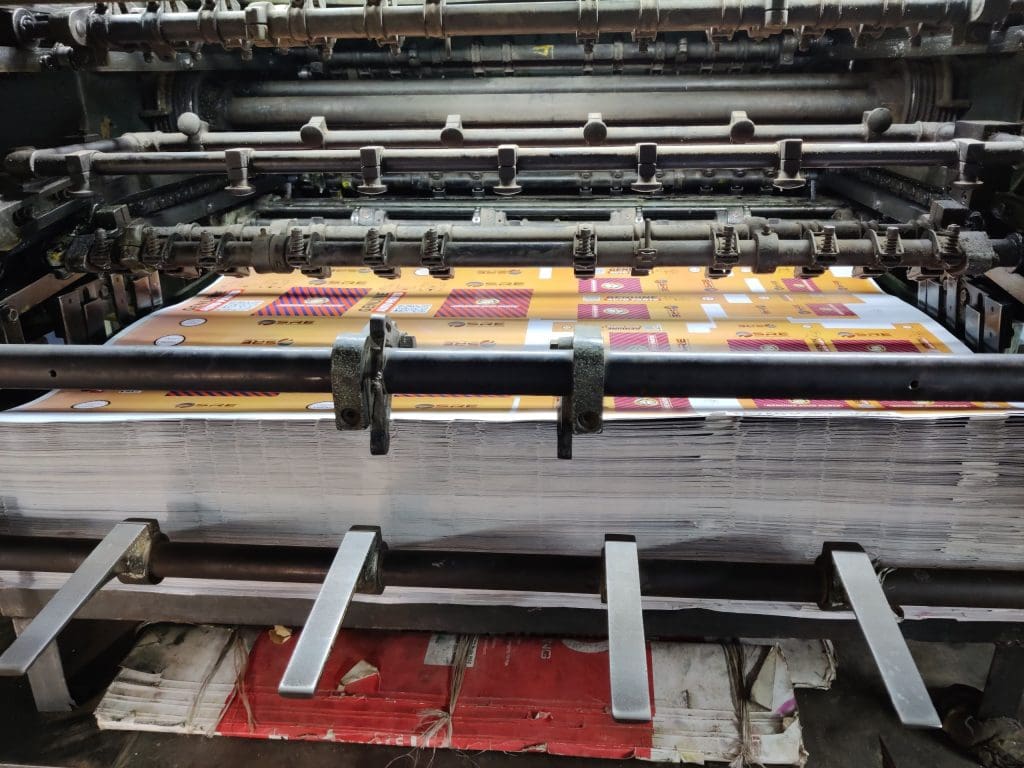 A close-up view of a printing press machine with a large stack of printed sheets inside. The sheets have a yellow and red design with text and graphics. The machine has multiple metal rods, gears, and clamps visible, showing the mechanical components involved in the printing process. The area beneath the machine has some worn and folded cardboard or paper materials.
